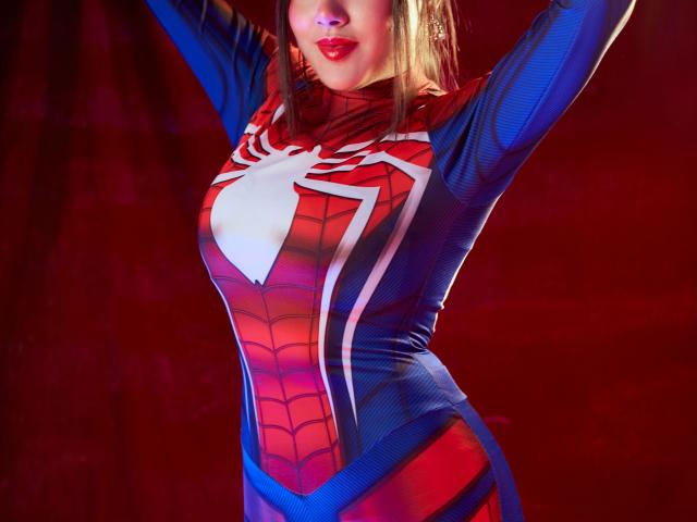 HannaGoez in a tight Spider-Man costume, arms raised, showing her big tits, red lips, and a white spider emblem on a red-blue bodysuit.