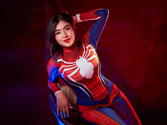 HannaGoez in a Spider-Man costume, red and blue with a white spider, has her hand on her head, sitting with a sexy smile, her ass slightly raised.