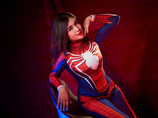 HannaGoez in a red and blue Spider-Man suit, posing with one hand on her head, showing off her Spider-Web chest, long dark hair, and a confident smile.