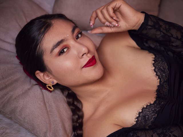 HannaGoez with dark hair, red lipstick, lace top, showing her tits, lying on a beige couch, wearing gold earrings, touching her nail, looking at the camera.