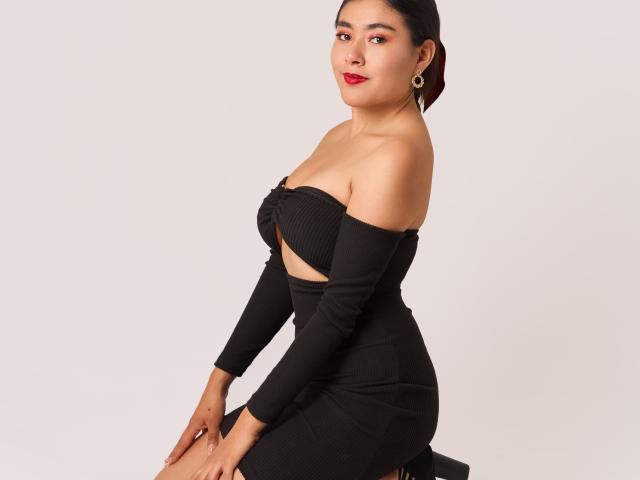 HannaGoez with dark hair, red lipstick, and gold earrings, wearing a black off-shoulder dress, sitting, showing her tits and ass in a tight, ribbed fabric.
