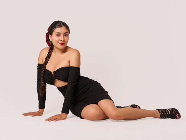 HannaGoez is sitting with her ass up, wearing a black off-shoulder dress, long braid, red lipstick, and black strappy heels, showing her tits and a cute smile.