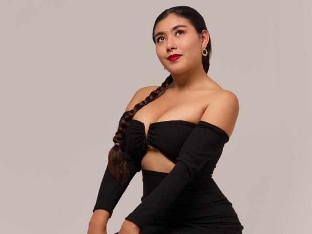 HannaGoez with dark braided hair, red lipstick, and hoop earrings, wearing a black off-shoulder top, exposing her tits and midriff, against a plain background.