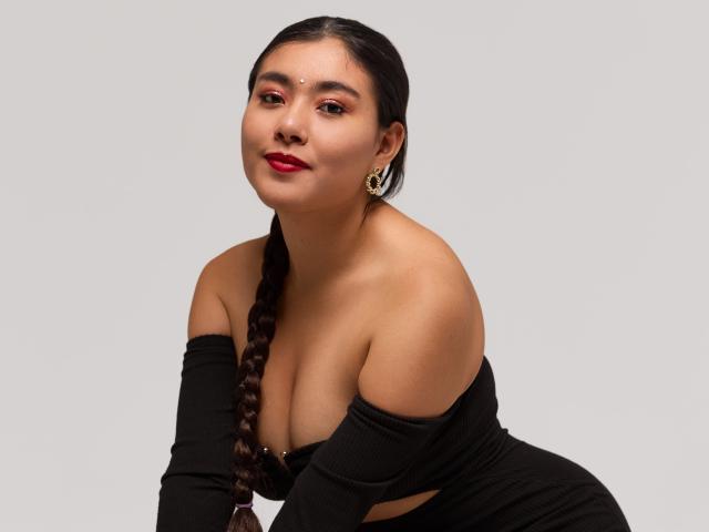 HannaGoez with dark braids, red lipstick, gold hoop earrings, black off-shoulder top, showing her big tits and bare shoulders, smiling slightly, on a plain gray background.