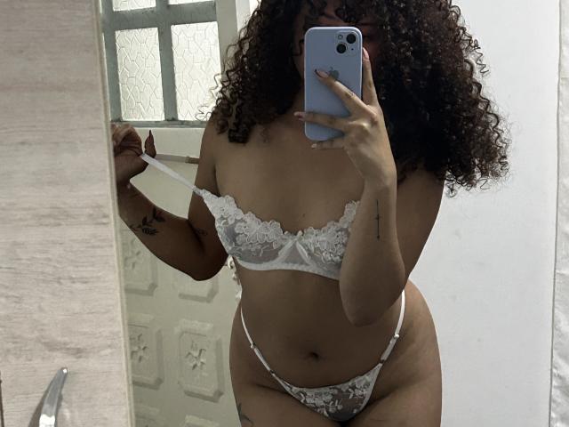 Sofia, passionnée, avec des cheveux bouclés, portant un soutien-gorge et une culotte en dentelle blanche, tenant un téléphone devant son visage, montrant ses seins et ses parties intimes dans un selfie pris dans un miroir..