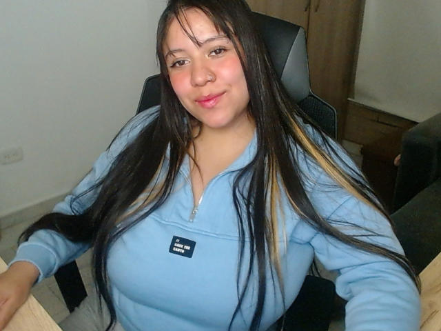 KrisGarner, com cabelos longos e pretos, mechas loiras e lábios rosados, vestindo um moletom azul com zíper, sentada em uma cadeira preta, exibindo seus seios grandes..