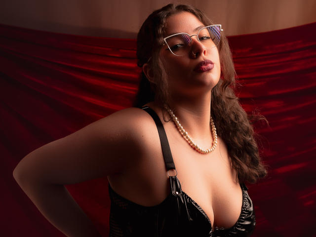 VanessaClose with glasses, pearl necklace, and black lace bra, showing off her big boobs and pouty lips, standing against red velvet. She's looking down with a confident expression.