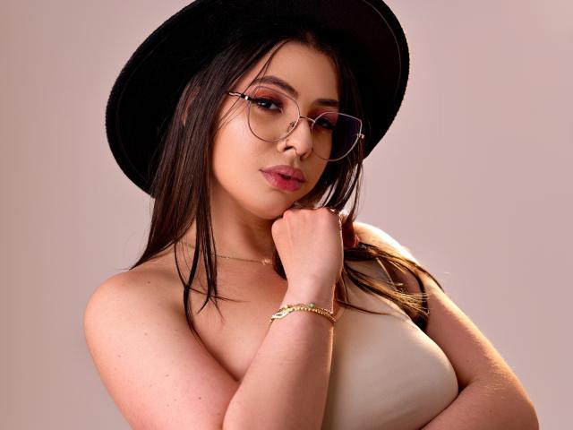 VanessaClose with glasses, black hat, and pink lipstick, touching her chin with her hand, wearing a gold bracelet, and a strapless white top, showing her tits.