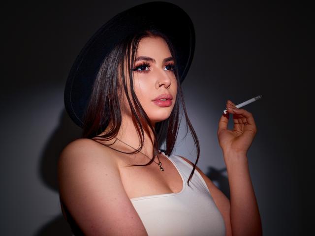 VanessaClose with dark hair, black hat, white top, holding a cigarette, brown eyes, pink lips, slightly parted, necklace with cross, shadowed background, confident, sexy, upper body.