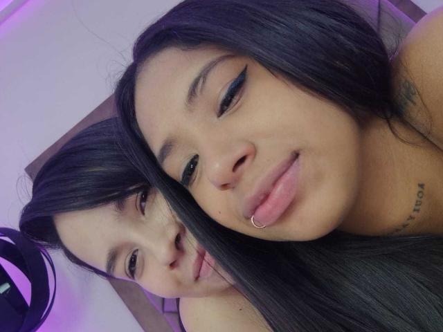 Dakota and Juliette, both with long black hair, winking and smiling. Dakota has a lip ring and a tattoo, while Juliette has a subtle smile in the background.