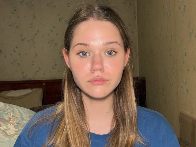 Juliet Erbst tiene ojos azules, piel clara y cabello castaño lacio. Lleva una camisa azul y tiene una expresión neutra, y se puede ver una pequeña peca en su mejilla derecha..