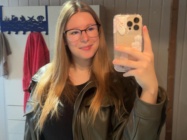 MadyN, met lang bruin haar, een bril en een zwarte leren jas, lacht in een spiegel-selfie en houdt een telefoon vast met een sticker van een wolk erop. Haar achterkant is niet zichtbaar..