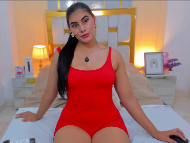 AlaraKey with long black hair, red dress, medium tits, and a necklace, sitting on a bed, looking at the camera, holding a black remote.