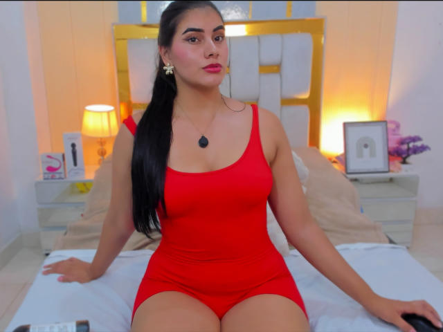AlaraKey with long black hair, red tank top, and red shorts, sitting on a bed, showing her tits, small black pendant, and pink lipstick.