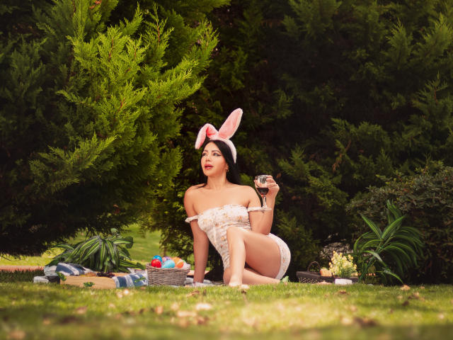 HelenHard with bunny ears, holding a glass, wearing a white off-shoulder top and shorts, sitting on grass with a picnic basket and Easter eggs, showing her ass.
