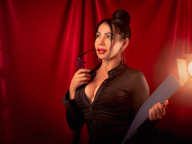 HelenHard with dark hair in a bun, wearing a ruched black shirt, holding glasses in one hand and a clipboard in the other, standing in front of red fabric.