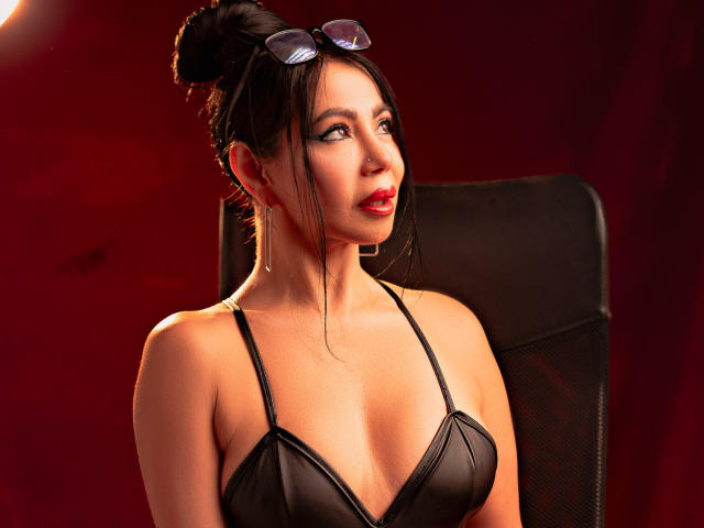 HelenHard with dark hair in a bun, wearing black leather bra, red lipstick, and glasses on head, looking to the side, sitting in black chair. Boobs visible.