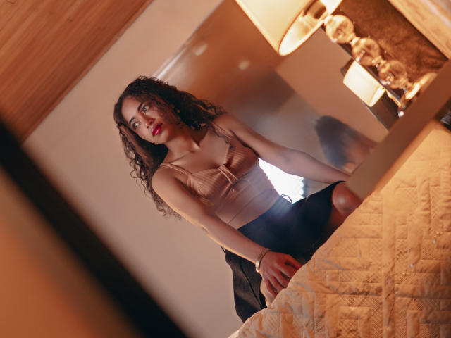 ViolettExposito with curly brown hair, red lipstick, beige top, and black skirt, leaning on bed, reflected in mirror, looking at camera, wood ceiling, warm lamp, quilted bedspread.