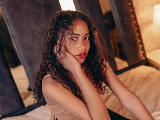 ViolettExposito with curly brown hair, dark skin, red lipstick, and a cute face, wearing a beige top, resting her chin on her hand, sitting on a bed.