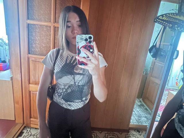 ShayWool with medium tits, wearing a graphic tee and black pants, taking a mirror selfie with a phone, wooden door in background, standing in a tiled room.