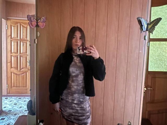 ShayWool, con i capelli castani, indossa una giacca nera e un abito tie-dye grigio, e tiene un telefono in un selfie allo specchio. Ci sono decorazioni a forma di farfalla sullo specchio. Il sedere di ShayWool è coperto..