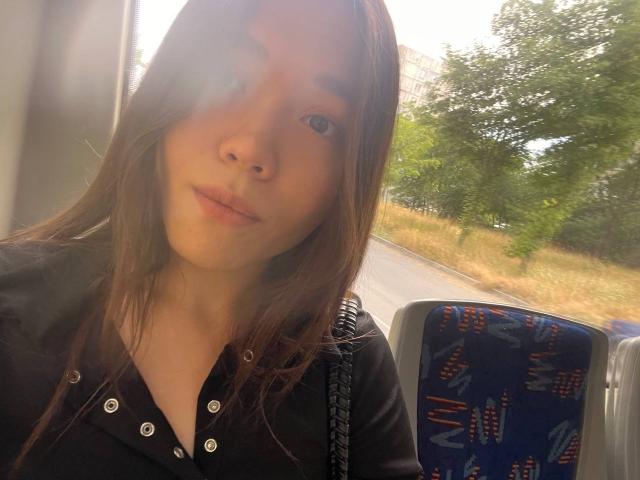 ShayWool has long brown hair, light skin, and is wearing a black button-up shirt, sitting in a bus with a blue patterned seat, looking slightly to the side.