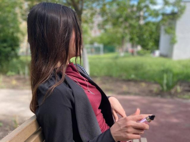 ShayWool with long brown hair, black jacket, red hoodie, holding a vape pen, sitting on a wooden bench, greenery in the background, blurred, daytime, relaxed, side profile.