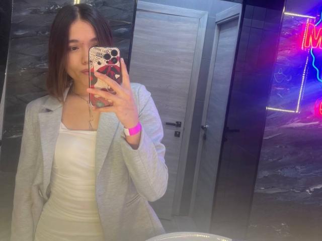 ShayWool avec une coupe de cheveux courte brune, portant un manteau gris par-dessus une robe blanche, tenant un téléphone avec une coque rose, éclairage néon, selfie devant un miroir, montre rose visible, poitrine de taille moyenne, mignonne, souriante..