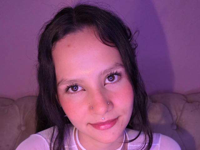 LilyVelle has dark, wavy hair, blue eyes, a small nose piercing, and a subtle smile with pinkish skin. She's wearing a white shirt, looking cute.