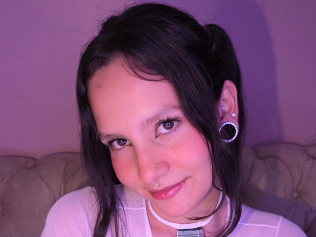 LilyVelle with black pigtails, blue eyes, nose piercing, lip piercing, white shirt, and silver hoop earrings, smiling softly. Purple light highlights her face.