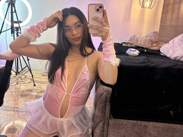 IsaDoux in rosa, indossa lingerie trasparente, con scollatura profonda, bordi bianchi, polsini vaporosi e una gonna corta, mentre si scatta un selfie con il telefono, mostrando i suoi seni e il suo piccolo sedere..
