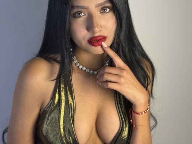 LillyTaylor with long black hair, red lipstick, and a shiny green halter top showing her big tits, touching her red lips with a finger, wearing a pearl necklace.