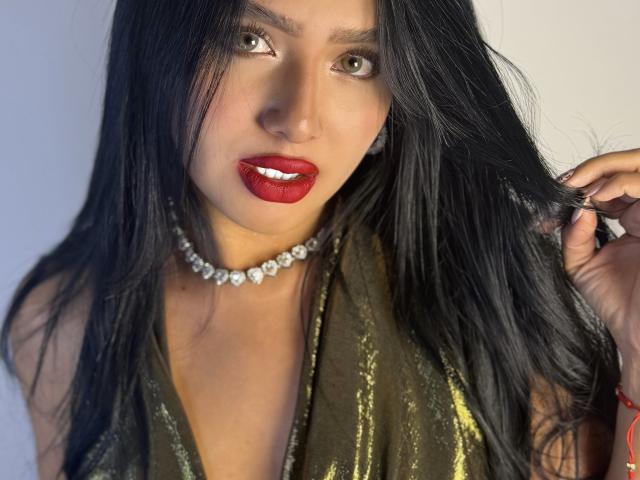 LillyTaylor with shiny black hair, red glossy lips, green eyes, wearing a sparkling choker and green shimmering dress, touching her hair. Boobs visible, no other visible body parts.