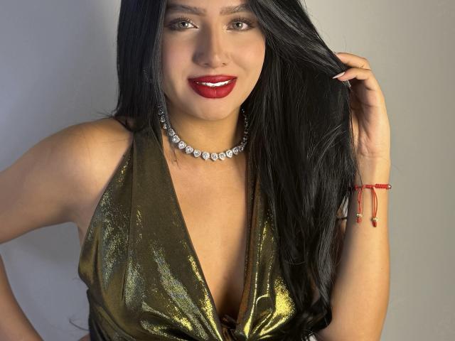 LillyTaylor with long black hair, red lipstick, green metallic dress, deep cleavage, diamond necklace, red bracelet, smiling, holding hair, sexy, tan skin, attractive.