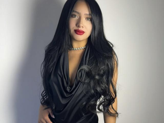 LillyTaylor with long black wavy hair, red lipstick, black sleeveless drape top, and diamond choker, looking at the camera with a slight smile. Her tits are small.