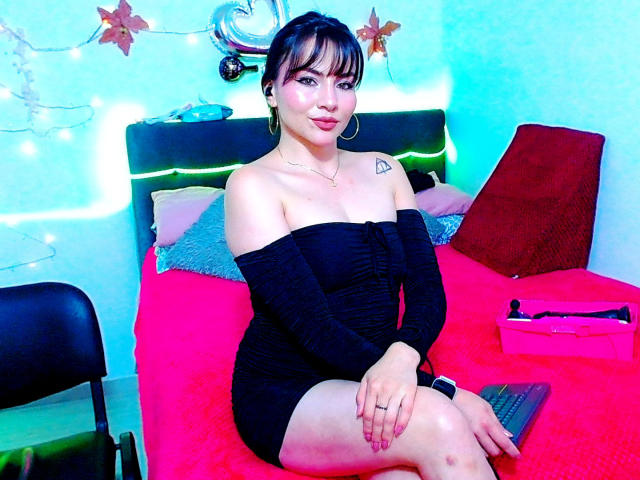 EmelySofia with black dress, off-shoulder, showing tits, small ass, tattoo on left shoulder, sitting on red bed, blue wall, chair, laptop, and open box with toys.