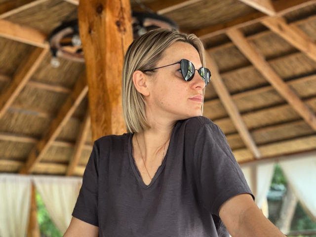 BelleLisaG with blonde bob, wearing aviator sunglasses and a dark V-neck tee, has a serious expression, wooden ceiling and rustic decor in the background.