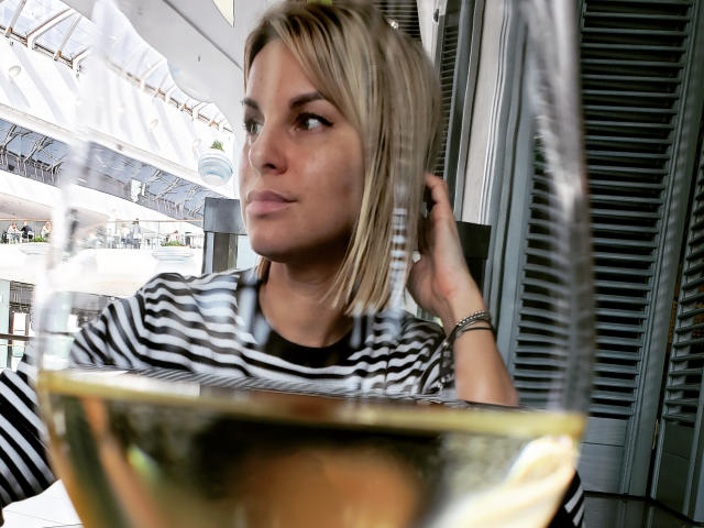 BelleLisaG with blonde bob, wearing black-and-white striped shirt, looking to the side, hand in hair, glass of white wine in foreground, blurred.