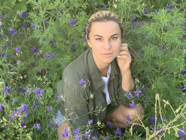 BelleLisaG with blonde hair, green eyes, and a serious look, wearing a green jacket, kneeling in a field of purple flowers, touching her ear with her right hand.