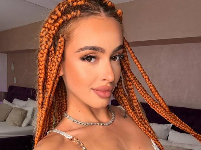MarissaFex with orange braided hair, full lips, and brown eyes, wearing a white strap top and diamond necklace, looking at the camera. Boob tattoo visible on her chest.