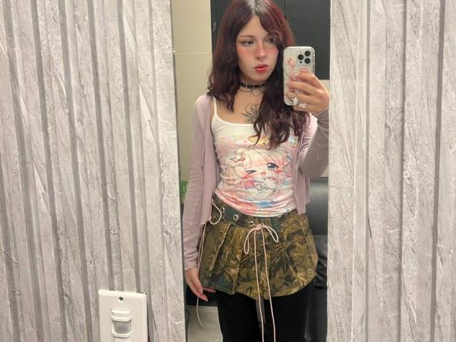 Valery Beaumont, com cabelos castanhos ondulados, vestindo uma camiseta rosa de anime, uma saia camuflada e um cardigã rosa, está tirando uma selfie no espelho com uma capa de celular estampada com flores. Seus seios são pequenos..