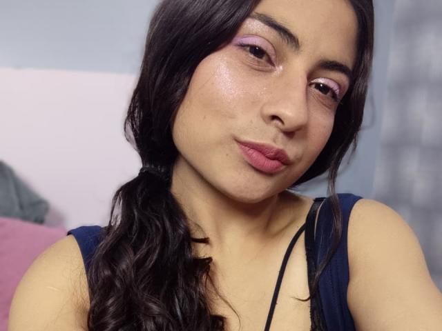 Angel Alba met opvallende roze lippenstift, glanzende huid en golvend donker haar in een staart, gekleed in een blauw topje, die haar volle, lipvormige billen laat zien in een selfie..