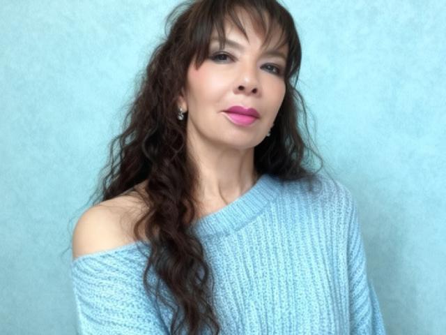 SamaraBellet, avec de longs cheveux bruns ondulés, du rouge à lèvres rose et un pull en tricot bleu clair à épaules dénudées, regarde légèrement vers la droite et porte de petites boucles d'oreilles..