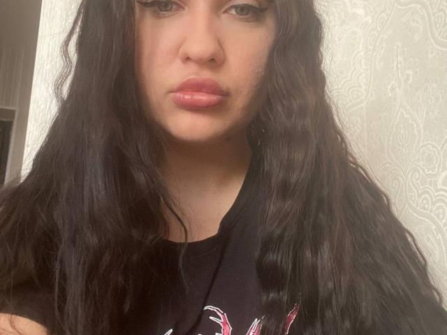 TroyJovanovic tem cabelos pretos longos e ondulados, lábios rosados cheios e olhos castanhos, vestindo uma camisa preta com um desenho rosa e branco. Seu rosto está centralizado na imagem..