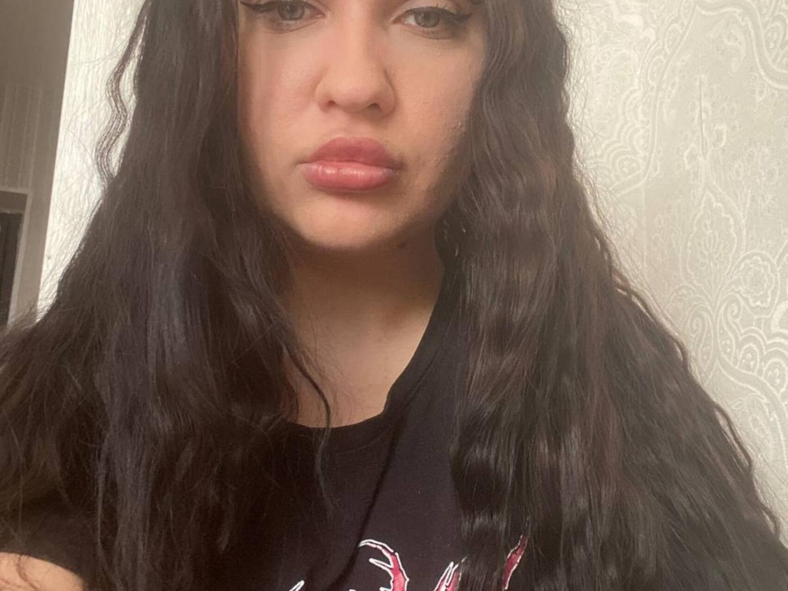 TroyJovanovic a de longs cheveux noirs ondulés, des lèvres pleines roses et des yeux marron, et elle porte une chemise noire avec un motif rose et blanc. Son visage est au centre de l'image..