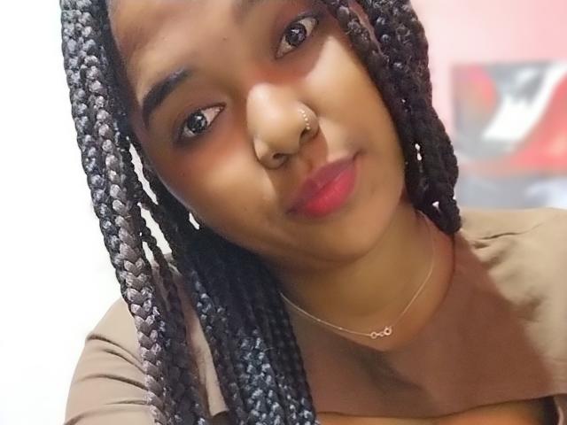 Shaniaha has dark braided hair, a nose ring, and wears a brown top with a gold necklace. Her pink lips are slightly parted in a subtle smile.