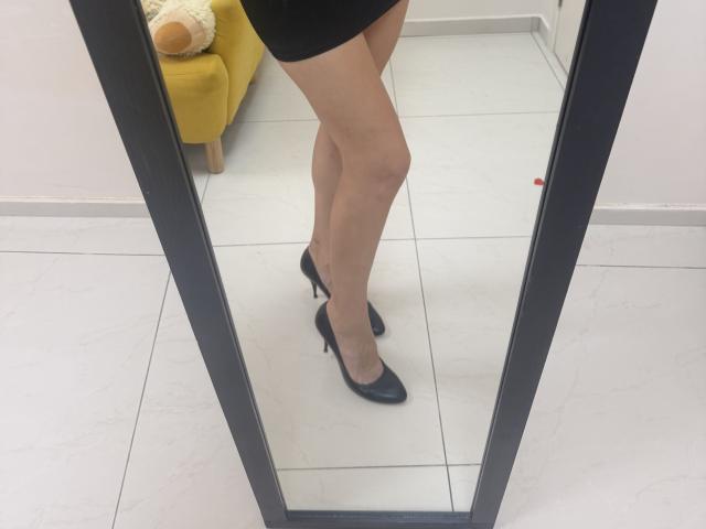 MoonLiza's bare legs in black high heels, reflected in a mirror, showing off her smooth, tan skin and cute ass. She's wearing a short black dress.