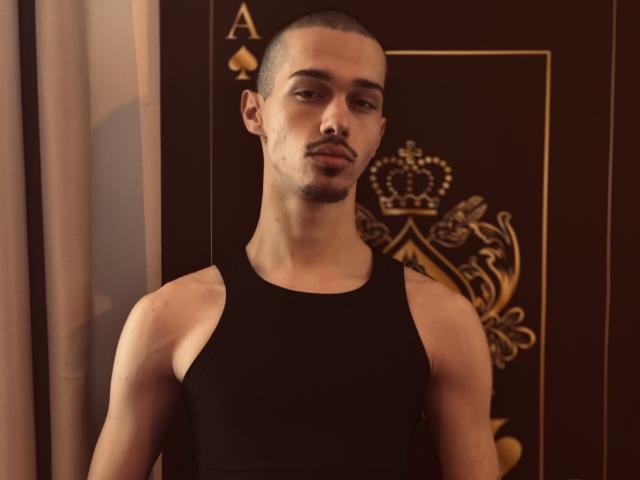 KaydenDrew tiene la cabeza rapada, piel marrón oscuro y una complexión delgada y musculosa. Lleva una camiseta de tirantes negra, que deja ver sus brazos y pecho tonificados..