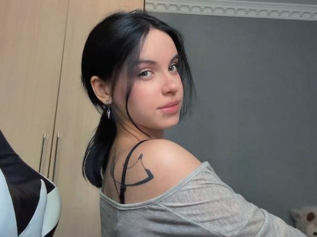 Rosenda Lazcano, con cabello negro, una blusa gris de corte asimétrico, mostrando un tatuaje negro en su hombro derecho, mirando por encima del hombro, sentada en una silla, con un piercing discreto en el labio..