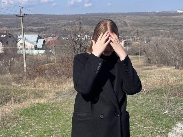 Tomiko Helfritz, com as mãos cobrindo o rosto, vestindo um casaco preto, em pé em um campo gramado, céu azul e nuvens esparsas, cabelo castanho, unhas vermelhas levemente visíveis..